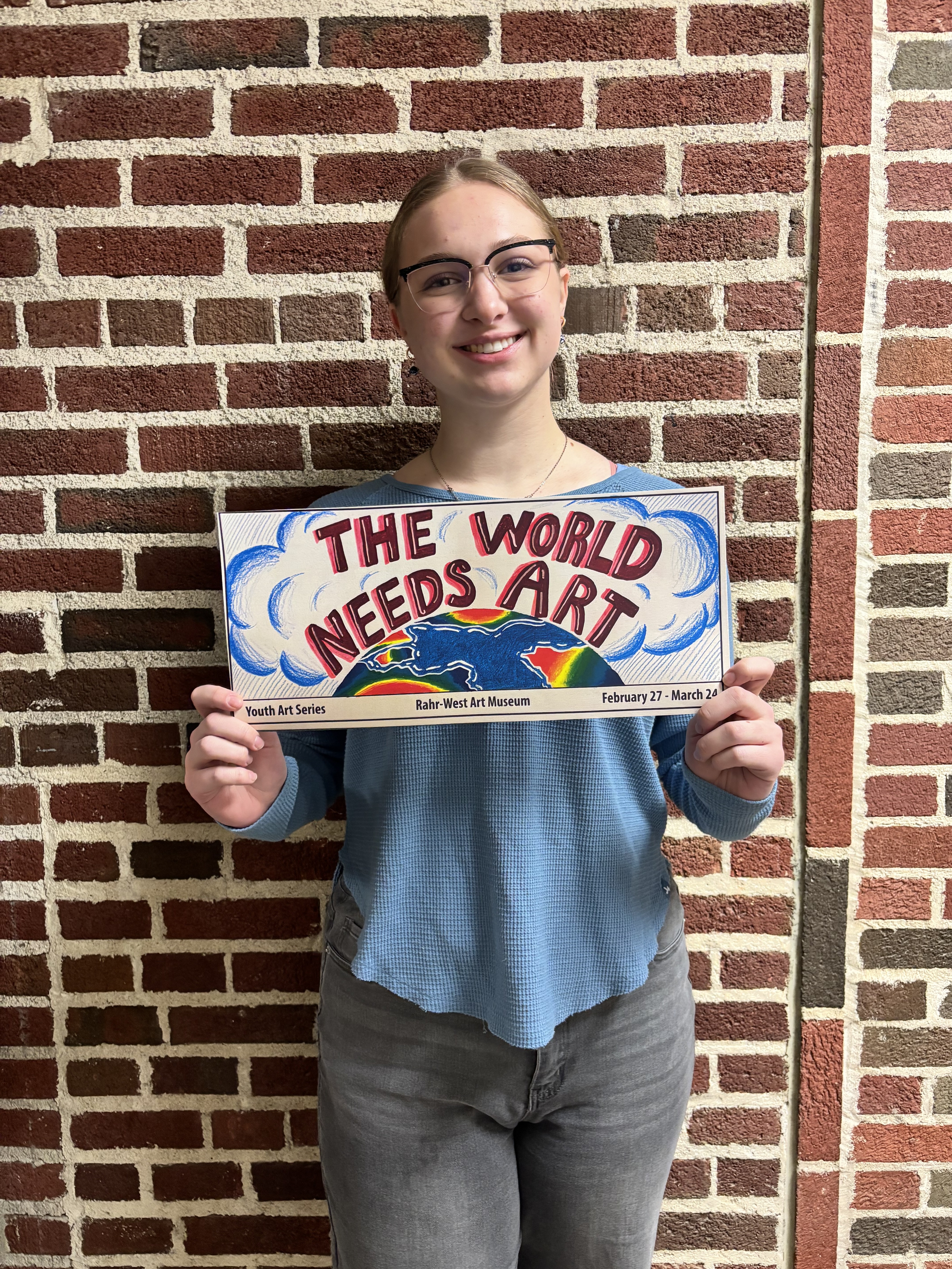 JACKIE DRAMM HOLDS BILLBOARD MOCKUP "THE WORLD NEEDS ART" ABOVE A COLORFUL GLOBE