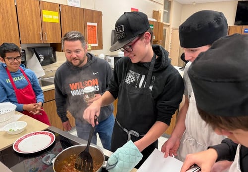 students cooking at a stove as their teacher watches on