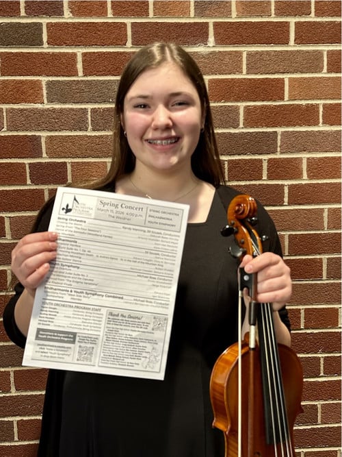 Soraya George stands with her violin and a Spring Concert pamphlet. She is in front of a brick wall.