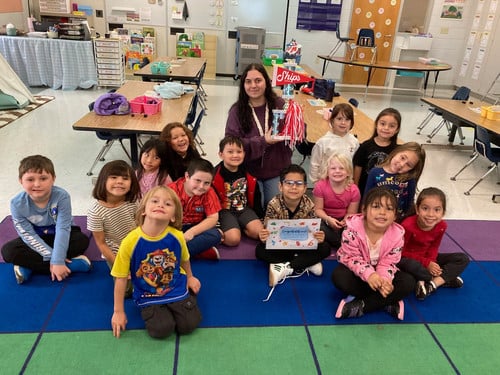 students sit on a carpet with their teacher who is holding an attendance trophy