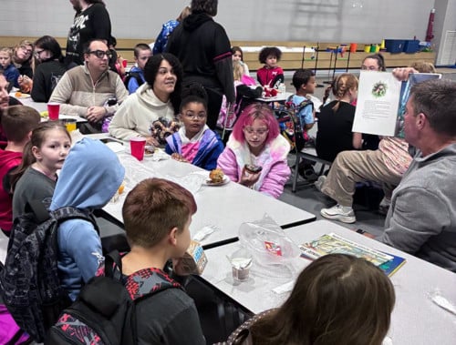 families sit at a cafeteria table with food listening to one man read a book while he shows the pictures
