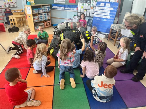 a firefighter kneels on the ground so students can feel how hard his helmet is