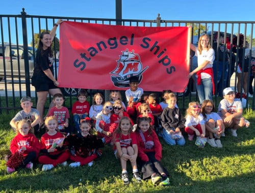 students sit in front of a stangel ships banner as teachers hold it for them
